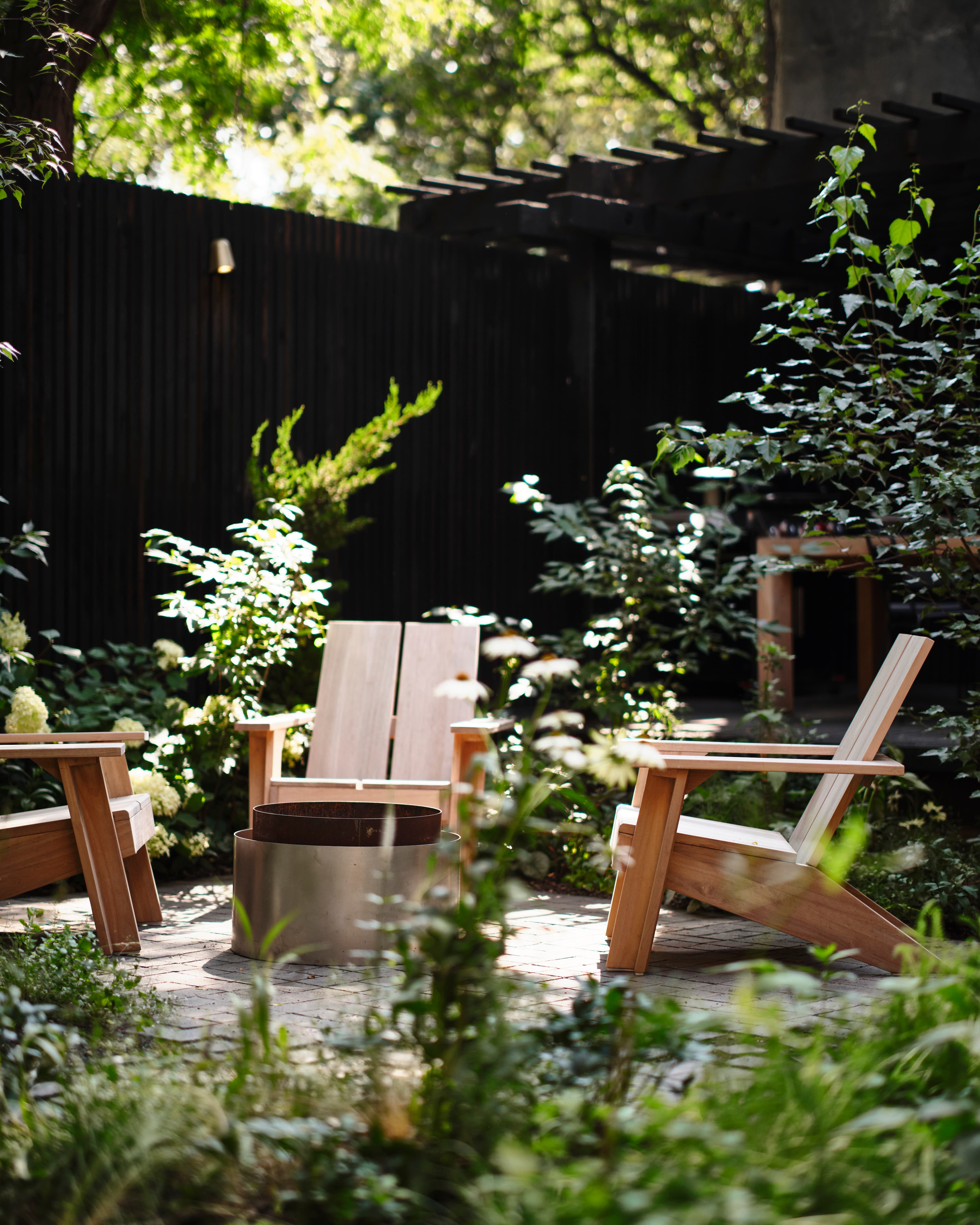 Blond-wood lounge chairs around a steel fire pit on basalt cobbles, nestled among hydrangea and juniper beneath a blackened pergola