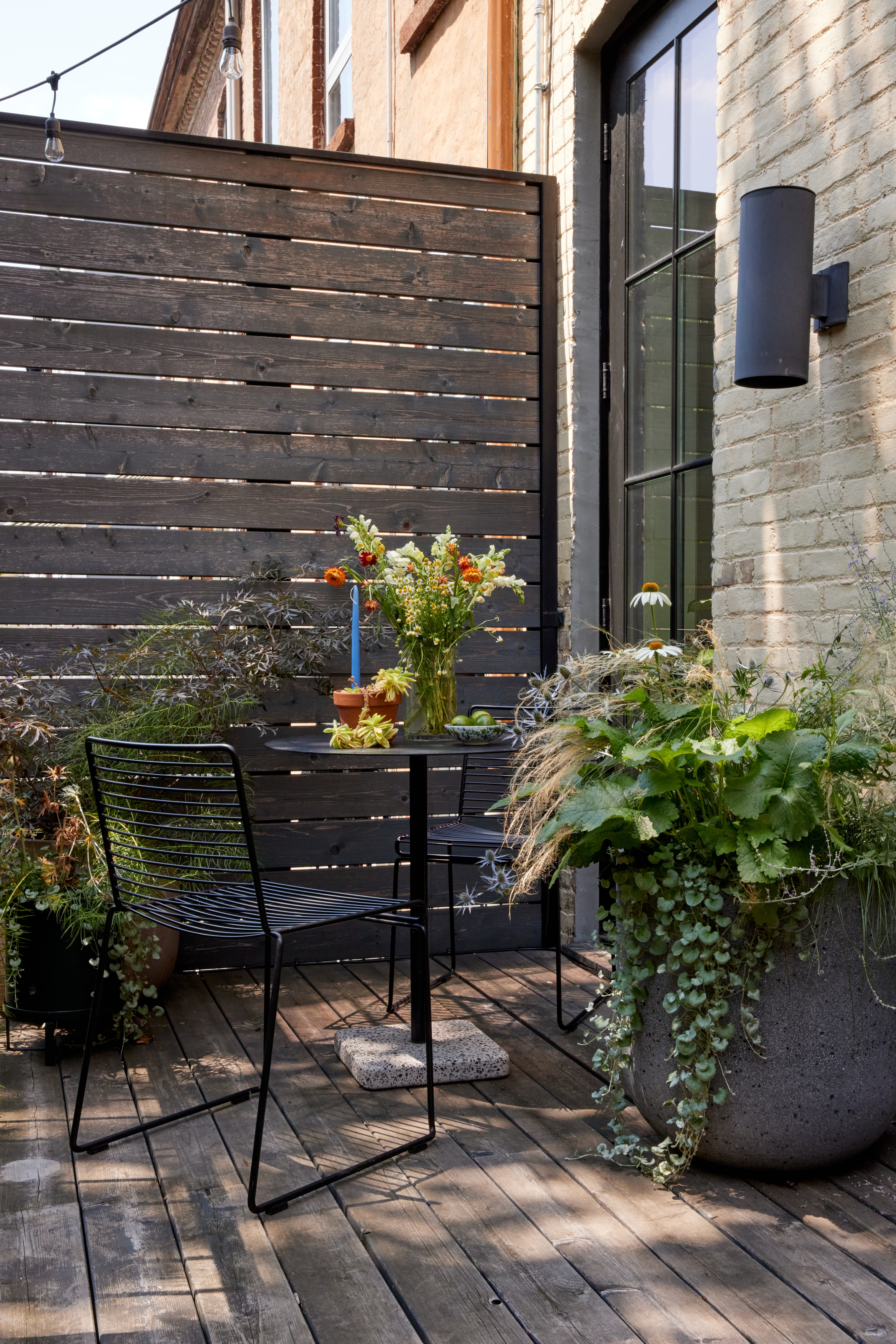 Deck bistro set against a slatted wood screen with black wire chairs, a Hay table, a cut-flower bouquet, and a concrete planter with trailing dichondra in a Brooklyn brownstone landscape