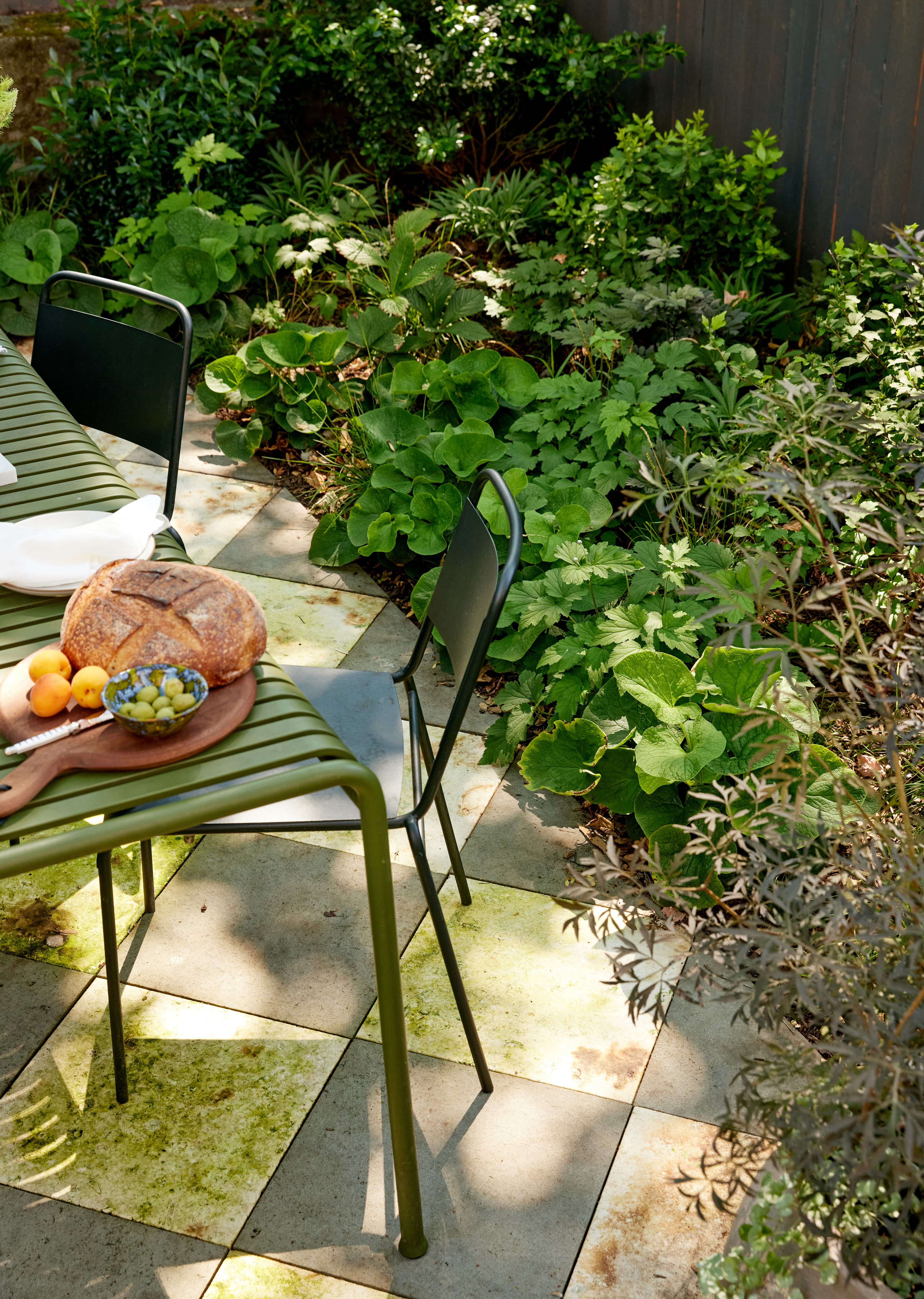 Alfresco table on checkerboard bluestone paving with crusty bread, apricots, and olives, edged by lush green shade planting in a New York landscape garden